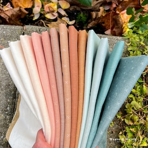 May include: A hand holds a fan of fabric swatches in various colors, including white, peach, coral, and teal. Each fabric has a subtle cross-stitch pattern. The swatches are arranged in a gradient, with the colors transitioning smoothly.
