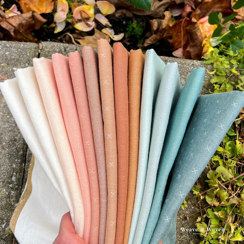May include: A hand holds a fan of fabric swatches in various colors, including white, peach, coral, and teal. Each fabric has a subtle cross-stitch pattern. The swatches are arranged in a gradient, with the colors transitioning smoothly.