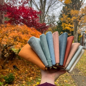 May include: A hand displays a selection of folded fabric squares in shades of orange, green, blue, and pink. Each square is decorated with a small white cross-stitch design. The background shows autumnal trees.
