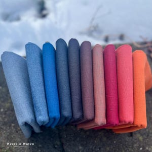 Peut inclure: Une collection de tissus pliés de différentes couleurs, notamment des nuances de bleu, de violet, de rose et d'orange. Le tissu est disposé en rangée, avec les bords visibles. Le texte "Weave & Woven" est en bas à gauche.