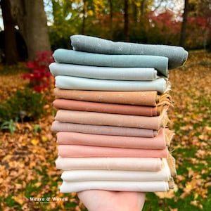 May include: A stack of folded fabric in various colors, including shades of blue, brown, and pink. Each piece has a subtle cross-stitch pattern. The fabric is held in a hand, with a blurred background of autumn leaves and trees.