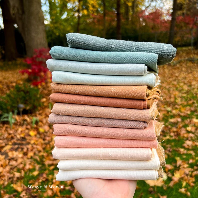May include: A stack of folded fabric in various colors, including shades of blue, brown, and pink. Each piece has a subtle cross-stitch pattern. The fabric is held in a hand, with a blurred background of autumn leaves and trees.