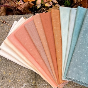 May include: A fan of various fabric swatches in shades of peach, orange, and blue. Each fabric has a subtle cross-stitch pattern. The fabrics are arranged on a stone surface, with some fall leaves in the background. The Weave & Woven logo is visible.