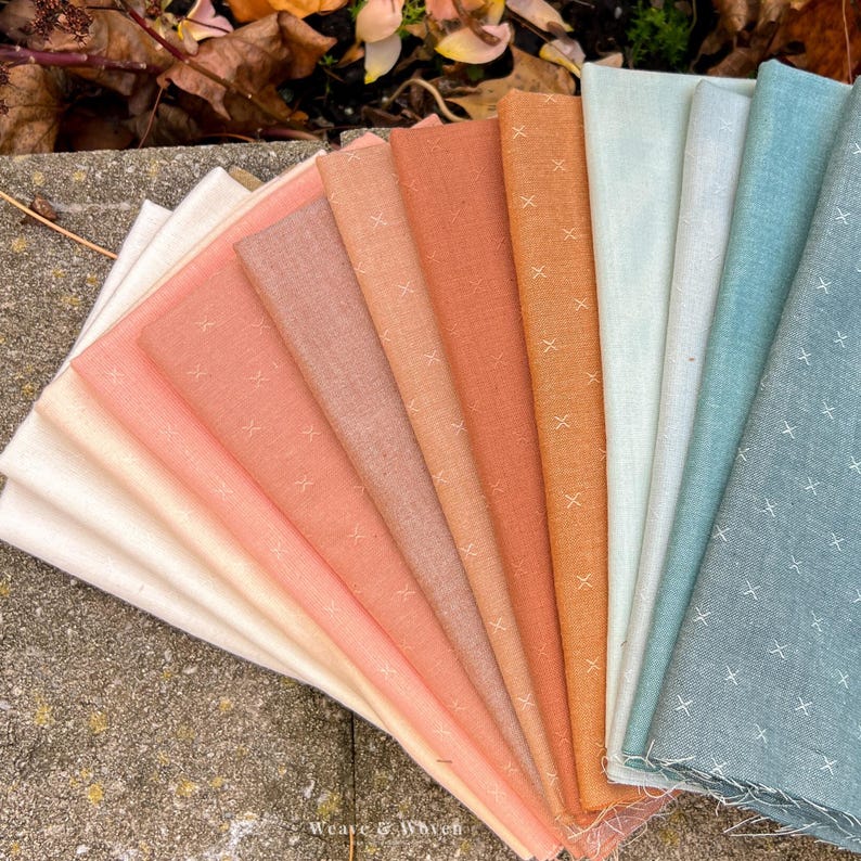 May include: A fan of various fabric swatches in shades of peach, orange, and blue. Each fabric has a subtle cross-stitch pattern. The fabrics are arranged on a stone surface, with some fall leaves in the background. The Weave & Woven logo is visible.