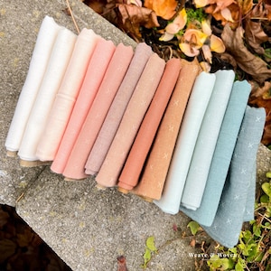May include: A collection of folded fabric squares in a variety of pastel colors, including white, pink, peach, and blue. Each square has a small embroidered cross pattern. The text "Weave & Woven" is visible in the bottom right corner.