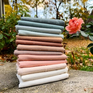 May include: A stack of folded fabric in various colors, including shades of green, brown, and pink. The fabric appears to be linen or a similar natural material. The top layers have a subtle cross-stitch pattern. The image is set outdoors with a rose in the background.