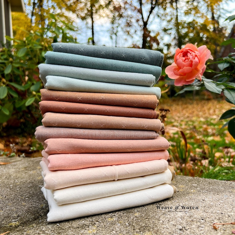 May include: A stack of folded fabric in various colors, including shades of green, brown, and pink. The fabric appears to be linen or a similar natural material. The top layers have a subtle cross-stitch pattern. The image is set outdoors with a rose in the background.