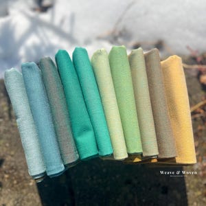 Peut inclure: Une collection de tissus pliés dans diverses nuances de vert, de turquoise et de jaune. Les tissus sont disposés en dégradé, les couleurs passant des tons froids aux tons chauds. L'image est bien éclairée, mettant en valeur la texture et les variations de couleur des textiles.