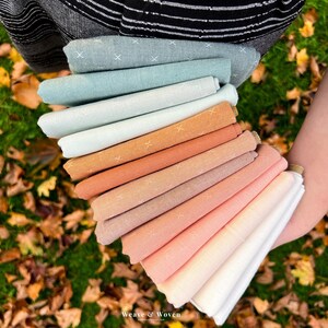 May include: A hand holds a stack of folded fabric in various pastel colors, including blue, orange, and pink. Each fabric piece has a subtle cross-stitch pattern. The fabrics are arranged in a fan shape, showcasing the range of hues.