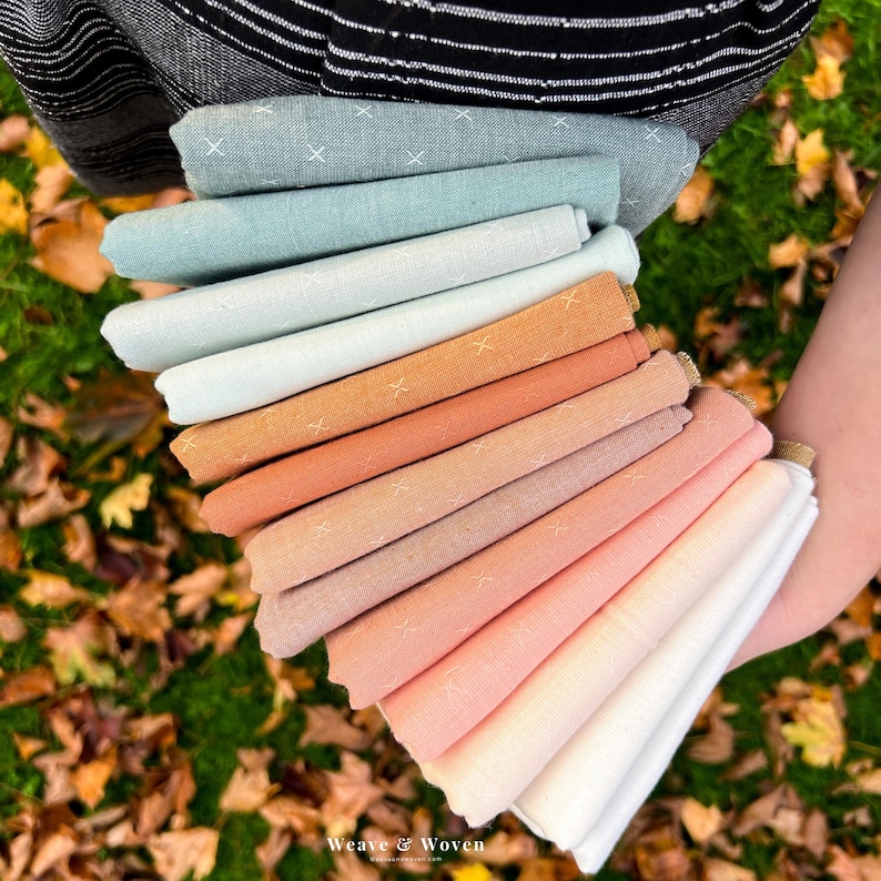 May include: A hand holds a stack of folded fabric in various pastel colors, including blue, orange, and pink. Each fabric piece has a subtle cross-stitch pattern. The fabrics are arranged in a fan shape, showcasing the range of hues.
