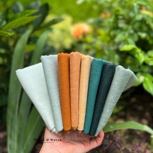 Peut inclure: Une main tient un éventail de serviettes en lin pliées de différentes couleurs, dont vert clair, turquoise, orange et beige. Les serviettes ont un aspect texturé et certaines présentent un motif de point de croix. L'arrière-plan est une vue floue de feuillage vert.