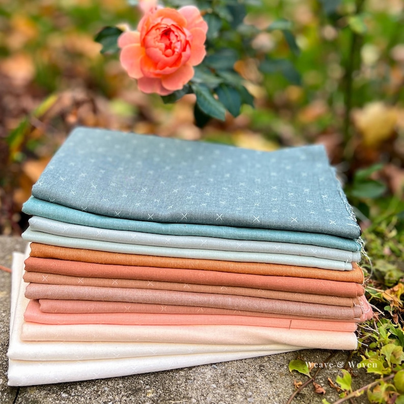 May include: A stack of fabric squares in various colors, including blue, green, orange, and pink. The fabrics have a subtle cross-stitch pattern. A rose in shades of peach and pink blooms in the background. The words "Weave & Woven" are in the bottom right corner.