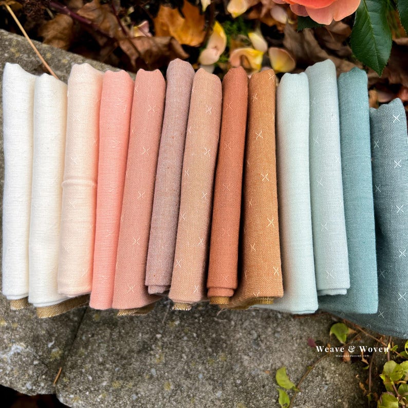 May include: A collection of folded fabric napkins in a variety of colors, including white, peach, and teal. Each napkin has a subtle cross-stitch pattern and a gold trim. The napkins are arranged in a row, showcasing their different hues.