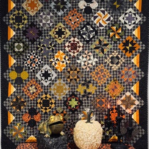 May include: A quilt featuring a grid of diamond-shaped blocks in orange, black, and grey patterns, displayed behind a table covered in an orange and black polka dot cloth, a green frog figurine on a rock, a white bumpy pumpkin, and a black candelabra.