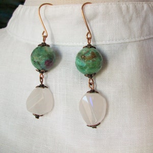 May include: A pair of dangle earrings with green and white gemstone beads. The green beads are round and have a mottled pattern. The white beads are teardrop shaped and have a translucent appearance. The earrings are made with copper wire and have a simple design.