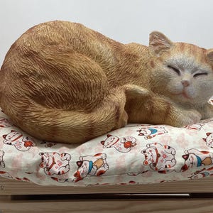 May include: A ginger cat figurine resting on a patterned pillow. The cat is curled up, eyes closed, on a pillow decorated with lucky cat illustrations. The pillow rests on a wooden crate.