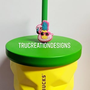 May include: A yellow Starbucks cup with a green lid and a pineapple-shaped straw topper with sunglasses. The straw topper is pink and blue with a white flamingo on top. The text "TRU CREATION DESIGNS" is on the lid.