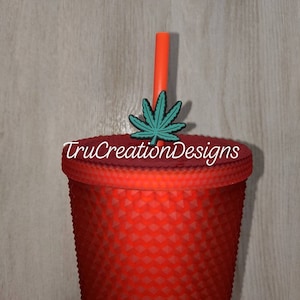 May include: Red tumbler with a textured surface, topped with a red lid and an orange straw. A teal cannabis leaf charm is attached to the lid. The text "TruCreationDesigns" is printed on the lid.