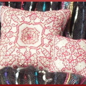 Puede incluir: Dos almohadas pequeñas, blancas, bordadas con hilo rojo. Las almohadas tienen un patrón floral y están sobre una tela de rayas blancas y negras.