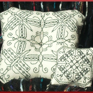 Puede incluir: Dos almohadas blancas con diseños geométricos bordados en negro. La almohada más grande tiene un diseño más intrincado con patrones repetidos de pájaros o insectos estilizados. La almohada más pequeña tiene un diseño más simple con un patrón repetido de tréboles de cuatro hojas.