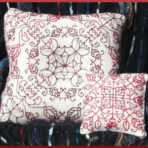 Puede incluir: Dos fundas de almohada blancas con bordados rojos. La almohada más grande tiene un diseño más intrincado con un patrón floral. La almohada más pequeña tiene un diseño geométrico más simple.
