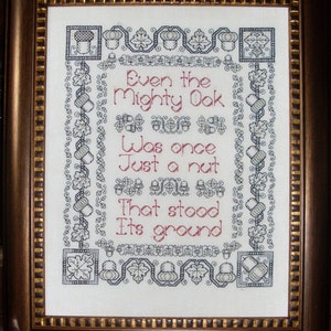 May include: A framed cross-stitch embroidery with a white background and black and gray stitching. The embroidery features a poem that reads "Even the mighty oak was once just a nut that stood its ground".