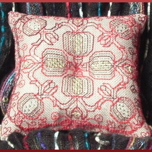 Puede incluir: Una pequeña almohada blanca con un diseño bordado en rojo y dorado de flores estilizadas. La almohada está sobre una tela oscura con estampado.