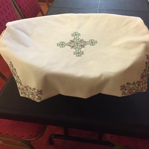 Puede incluir: Mantel blanco con un borde floral y una gran cruz bordada en el centro. La cruz es verde, rosa y amarilla.