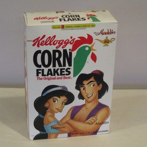 Op de afbeelding: Een witte ontbijtgranen doos met Kellogg's Corn Flakes, een rode haan en een groene achtergrond. De doos heeft een Disney Aladdin-thema met afbeeldingen van Aladdin en Jasmine. De tekst op de doos luidt "Kellogg's Corn Flakes The Original and Best."