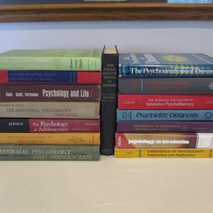 May include: A stack of vintage psychology books with titles like "Psychology and Life" and "The Psychoanalysis of Dreams." The books are in various colors, including green, red, and blue, and are arranged on a light-colored surface.