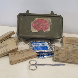 May include: An olive green metal first aid kit with a faded red and white label. The kit is open, revealing various medical supplies, including bandages, gauze, and Vaseline. A pair of silver scissors rests in front of the kit.