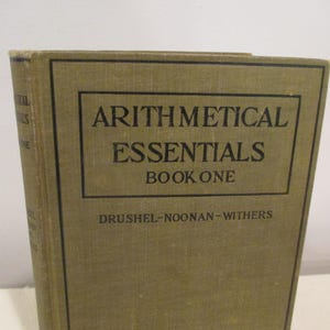 May include: A vintage book titled "Arithmetical Essentials Book One" by Drushel-Noonan-Withers. The book has a brown linen cover with black text.