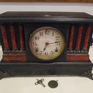 Puede incluir: Reloj de chimenea negro antiguo con detalles rojos y dorados. El reloj tiene una esfera redonda con números romanos y un bisel dorado. Incluye una llave y una pequeña pieza metálica redonda.