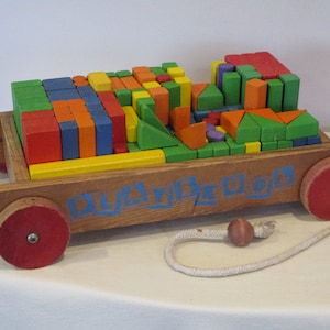 Könnte beinhalten: Ein hölzerner Spielzeugwagen gefüllt mit bunten Holzwürfeln. Der Wagen hat rote Räder und ein blaues Seil, das daran befestigt ist. Der Wagen ist mit "PLAYSKOOL" beschriftet.
