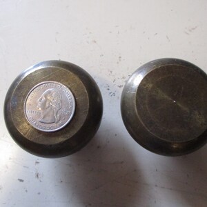Puede incluir: Dos pomos de latón antiguos. Uno tiene una moneda de veinticinco centavos para comparar el tamaño. Los pomos son redondos con una parte superior plana y un cuerpo ligeramente abovedado. El latón tiene una pátina cálida y envejecida.