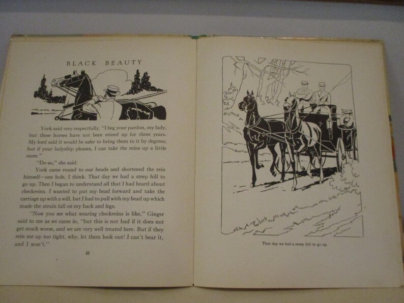 May include: An open book with illustrations from "Black Beauty." The left page features a drawing of a horse-drawn carriage and text. The right page shows a detailed illustration of a horse-drawn carriage on a hill. The book is open on a light-colored surface.