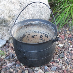 Może przedstawiać: Czarny kocioł żeliwny z metalowym uchwytem. Kocioł stoi na warstwie małych kamieni.