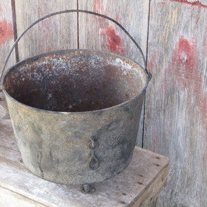 Puede incluir: Una olla grande de hierro fundido oxidada con un asa de alambre y tres patas. La olla está sentada sobre una superficie de madera.