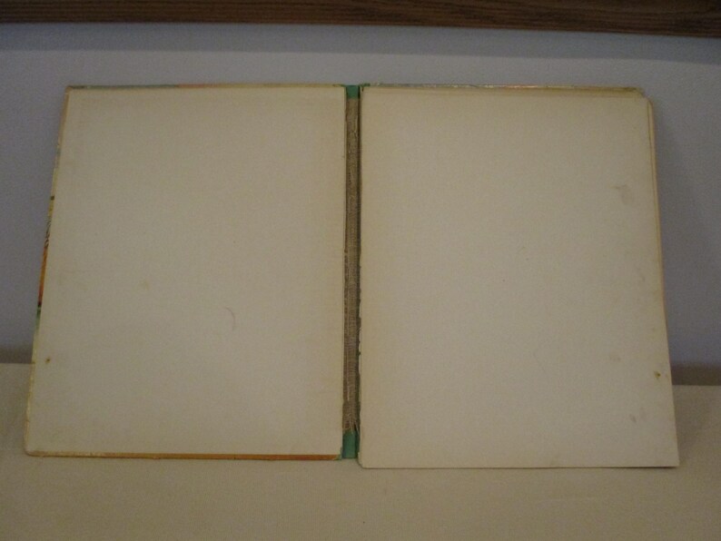 May include: An open vintage book with off-white pages. The book's spine is visible, with a green and tan binding. The pages show slight discoloration, suggesting age. The book is set against a neutral background, highlighting its aged appearance.
