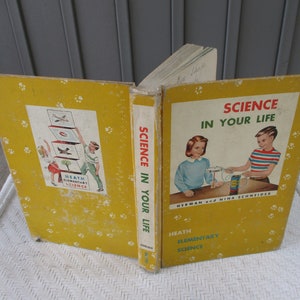 Könnte beinhalten: Ein gelbes Buch mit dem Titel "Wissenschaft in Ihrem Leben" und dem Untertitel "Heath Elementary Science". Der Umschlag zeigt Illustrationen von Kindern, die wissenschaftliche Experimente durchführen.