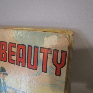 May include: Close-up of a vintage book cover with the title "Black Beauty" in large red letters with a black outline. The cover features an illustration of a person in a blue coat and hat. The book has a worn, aged appearance.