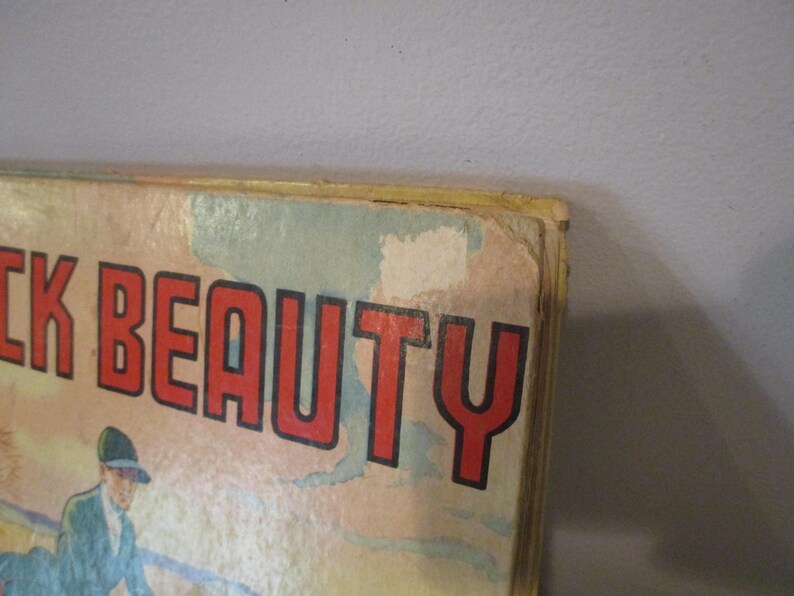 May include: Close-up of a vintage book cover with the title "Black Beauty" in large red letters with a black outline. The cover features an illustration of a person in a blue coat and hat. The book has a worn, aged appearance.