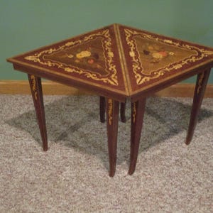 May include: A triangular wooden side table featuring intricate floral marquetry inlay on the top surface. The table has four slender, tapered legs with decorative gold-toned accents and a dark brown polished wood finish throughout.