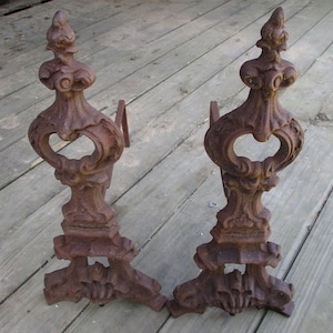May include: A pair of ornate cast iron fireplace andirons with a rust-colored finish. The andirons have a decorative design with a heart-shaped opening in the center.