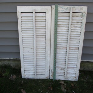 May include: Two weathered, white wooden shutters with horizontal slats and vertical supports. The shutters are leaning against a gray wall. A green ruler is placed between the shutters. The shutters show signs of age and wear.