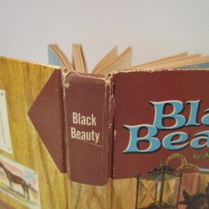 May include: An open book with the title "Black Beauty" by Anna Sewell. The cover is a deep red with blue lettering. The spine of the book is brown with the title printed vertically. The book is displayed with a wooden background.
