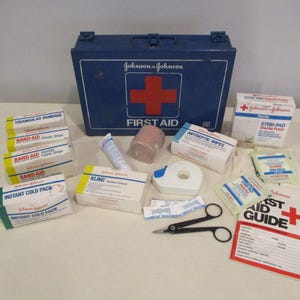 Puede incluir: Un botiquín azul de primeros auxilios Johnson & Johnson con una cruz roja, rodeado de vendas, toallitas antisépticas y una compresa fría instantánea. También se incluyen unas tijeras negras y una guía de primeros auxilios.