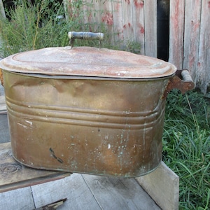 May include: A large, oval-shaped copper wash tub with a lid and two handles. The tub is rusty and has a patina. The tub is sitting on a wooden surface.