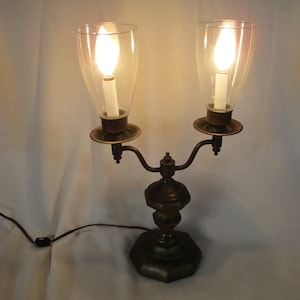 Puede incluir: Una lámpara de candelabro de latón vintage con dos pantallas de vidrio y bombillas. La lámpara tiene una base decorativa y un cable con enchufe.