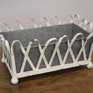 Op de afbeelding: Een rechthoekige metalen mand met een verweerde witte afwerking en een grijze binnenkant. De mand heeft een decoratief kruislings latwerkpatroon langs de zijkanten en rust op vier kleine, afgeronde, bolvormige voetjes op de hoeken.